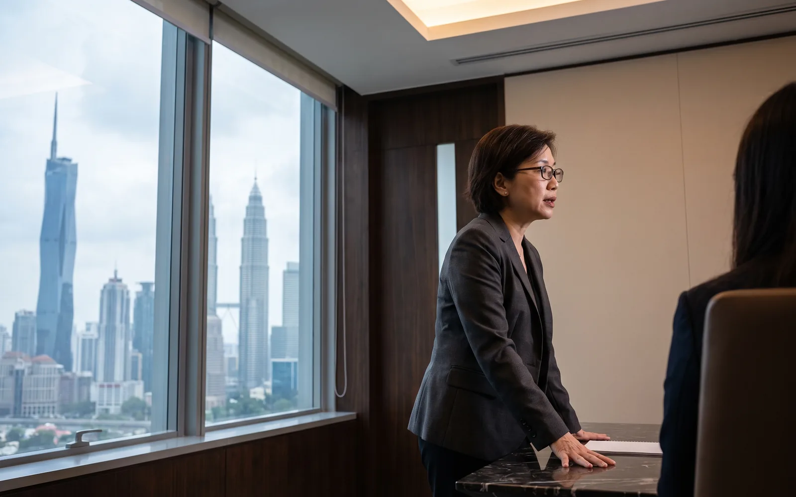 A senior executive in a calm, light-filled Kuala Lumpur office, the business district beyond the glass